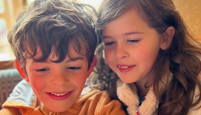 Two children sitting closely together indoors, with one wearing an orange zip-up jacket and the other in a white furry gilet over a blue shirt. The setting appears casual and warm. They are looking at a tablet or device partially visible in the foreground
