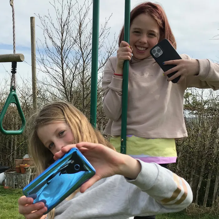 Two children outdoors on playground equipment, each holding a smartphone and appearing to take photos or videos. Background includes leafless trees and a cloudy sky, with green metal bars and a triangular climbing handle visible