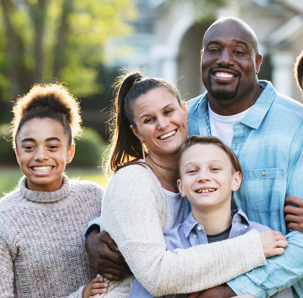A family of five standing outdoors in a suburban neighbourhood, with blurred trees and houses visible in the background. Family members are dressed casually and closely embracing, conveying a sense of warmth and togetherness.