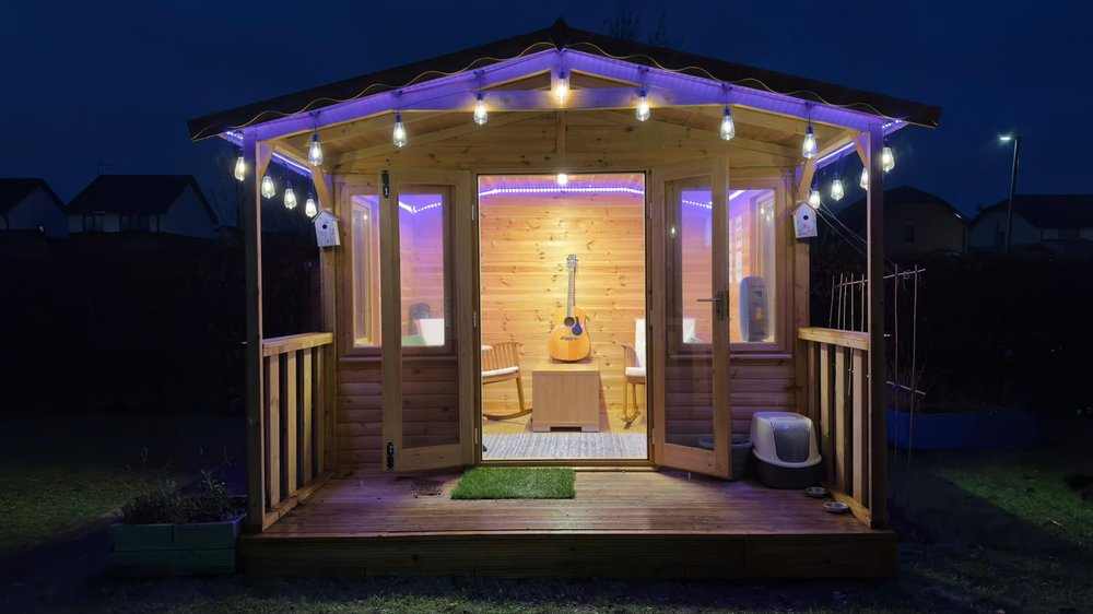 A small wooden garden shed glows with warm yellow and purple lights at night. The double doors are open, showing a guitar and two chairs inside. On the porch, there’s a cat litter box and food bowl. The scene feels cozy and welcoming.