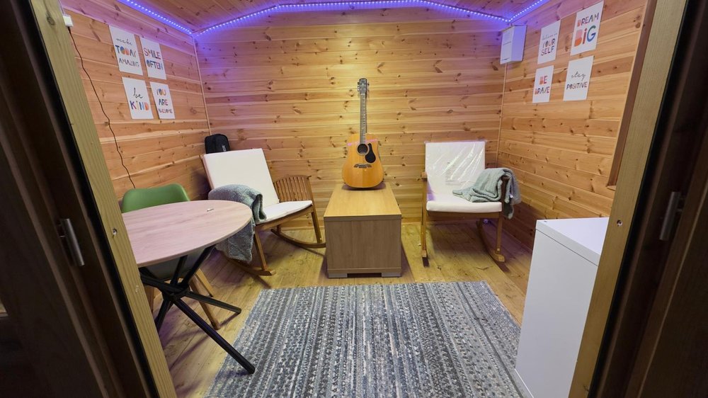 A warm, wooden room designed for comfort and creativity. Soft purple lights glow along the ceiling, and two cushioned rocking chairs sit facing a small wooden table with an acoustic guitar resting on top. A patterned rug covers part of the floor.