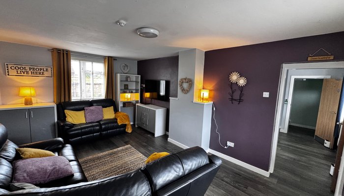 Tan y Dre's cosy living room. The walls are painted dark purple, with two leather sofas and a sign that says 'Cool people live here'