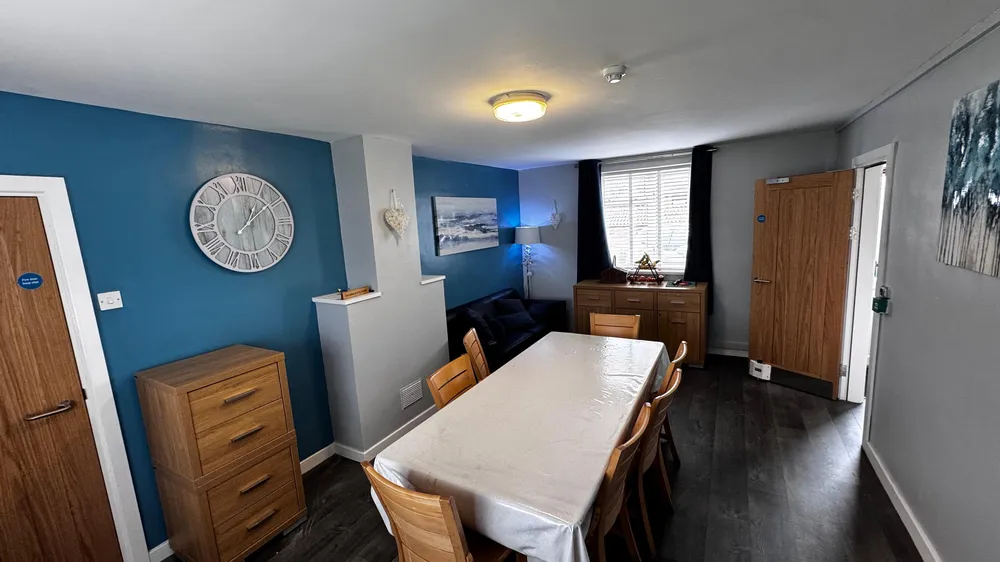 A modern dining and living area with a rectangular table covered by a white tablecloth, surrounded by wooden chairs. The room features blue and gray walls, a small sofa, decorative artwork, a large wall clock, and a window with dark curtains.