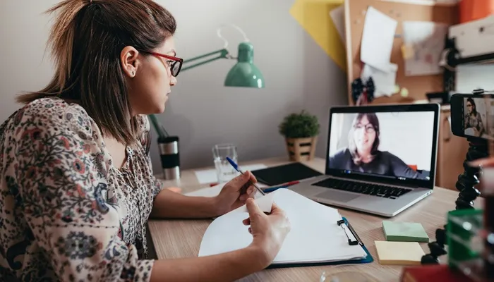A woman participating in a video call on a laptop while taking notes on a clipboard at a desk. The workspace includes a green desk lamp, sticky notes, a plant, and various office supplies, indicating a remote work or study environment.
