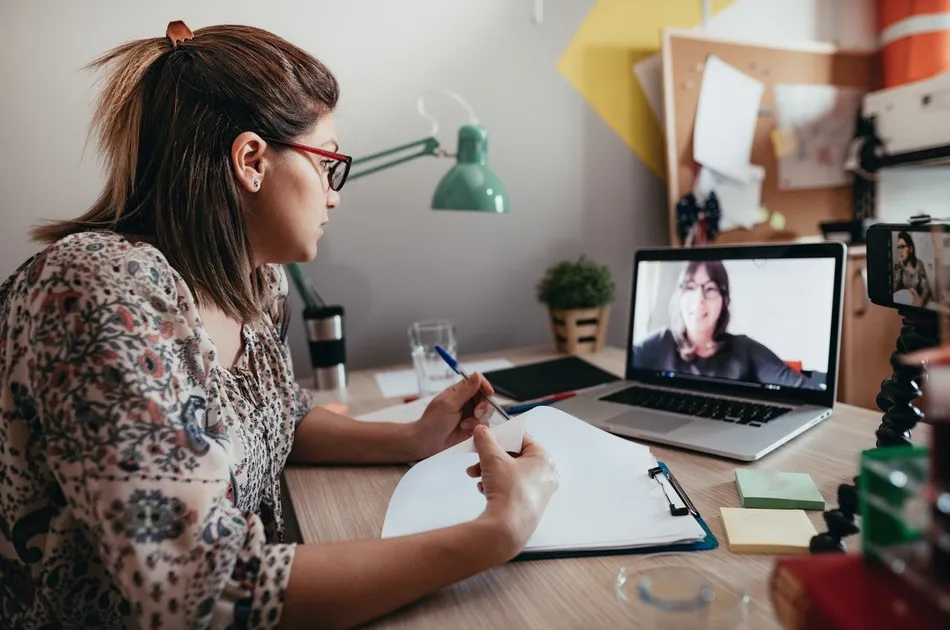 A woman participating in a video call on a laptop while taking notes on a clipboard at a desk. The workspace includes a green desk lamp, sticky notes, a plant, and various office supplies, indicating a remote work or study environment.