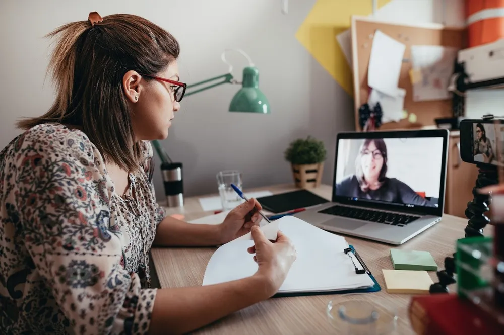 A woman participating in a video call on a laptop while taking notes on a clipboard at a desk. The workspace includes a green desk lamp, sticky notes, a plant, and various office supplies, indicating a remote work or study environment.