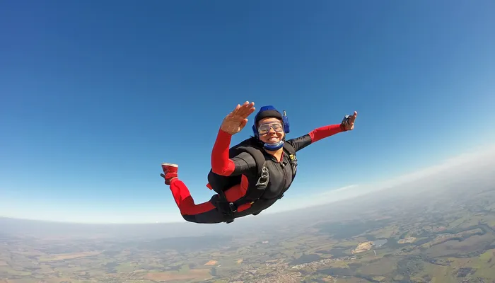 A smiling person skydiving above a rural landscape with clear blue sky. The skydiver wears a black jumpsuit with red sleeves and legs, a blue helmet, and is extending arms and legs in freefall position.