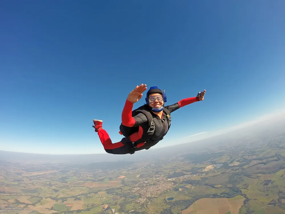 A smiling person skydiving above a rural landscape with clear blue sky. The skydiver wears a black jumpsuit with red sleeves and legs, a blue helmet, and is extending arms and legs in freefall position.