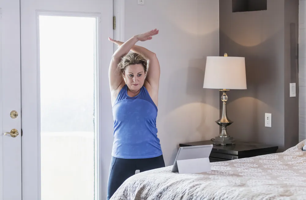 A person performing an overhead arm stretch in a bedroom setting, wearing a blue sleeveless top and black joggers. The scene includes a bed with a patterned bedspread, a bedside table with a lamp, and a tablet device, suggesting an online exercise class