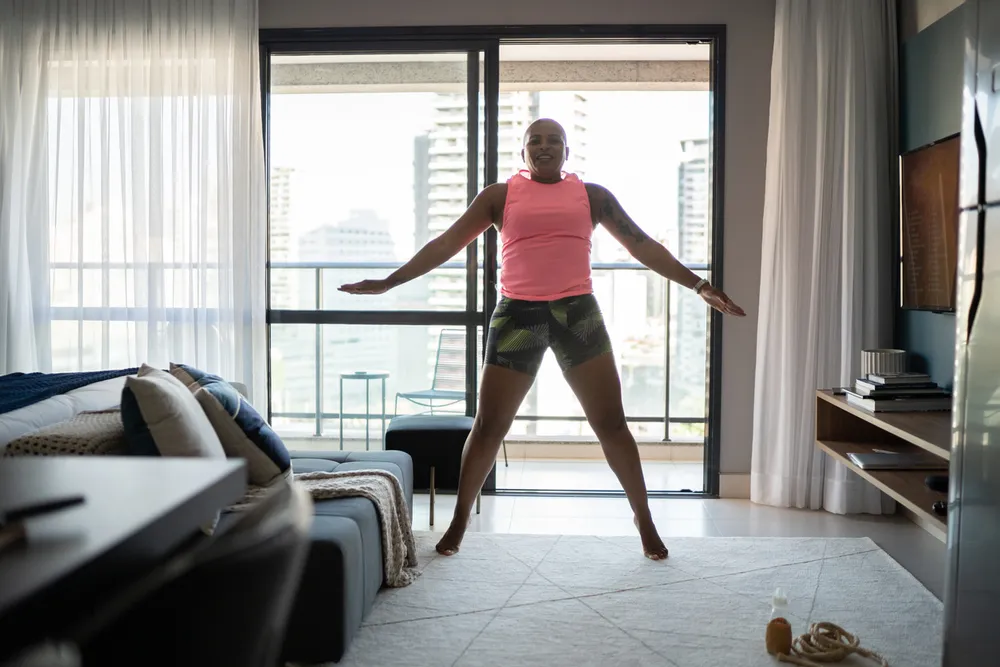 Woman is in her living room. She is wearing shorts and a tank top doing star jumps