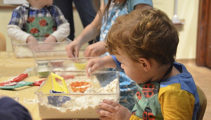 Young boy sat at table playing with a plastic box filled with sand and small colourful toys. More children playing in the background