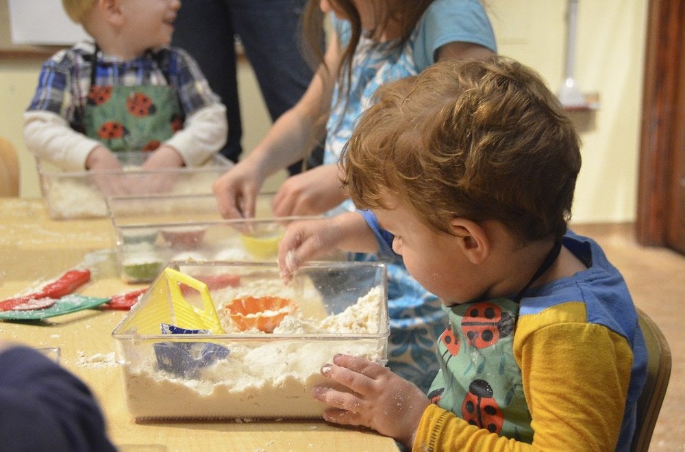 Young boy sat at table playing with a plastic box filled with sand and small colourful toys. More children playing in the background