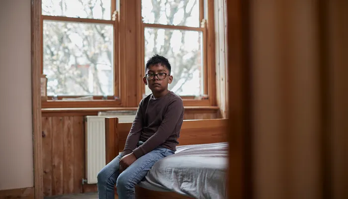A young boy sitting on the edge of a wooden bed in a sparsely furnished room with large windows showing bare tree branches outside. The scene conveys a quiet, contemplative mood. The boy is dressed in a brown long-sleeve top and blue jeans.