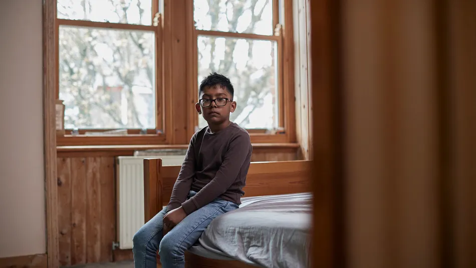A young boy sitting on the edge of a wooden bed in a sparsely furnished room with large windows showing bare tree branches outside. The scene conveys a quiet, contemplative mood. The boy is dressed in a brown long-sleeve top and blue jeans.