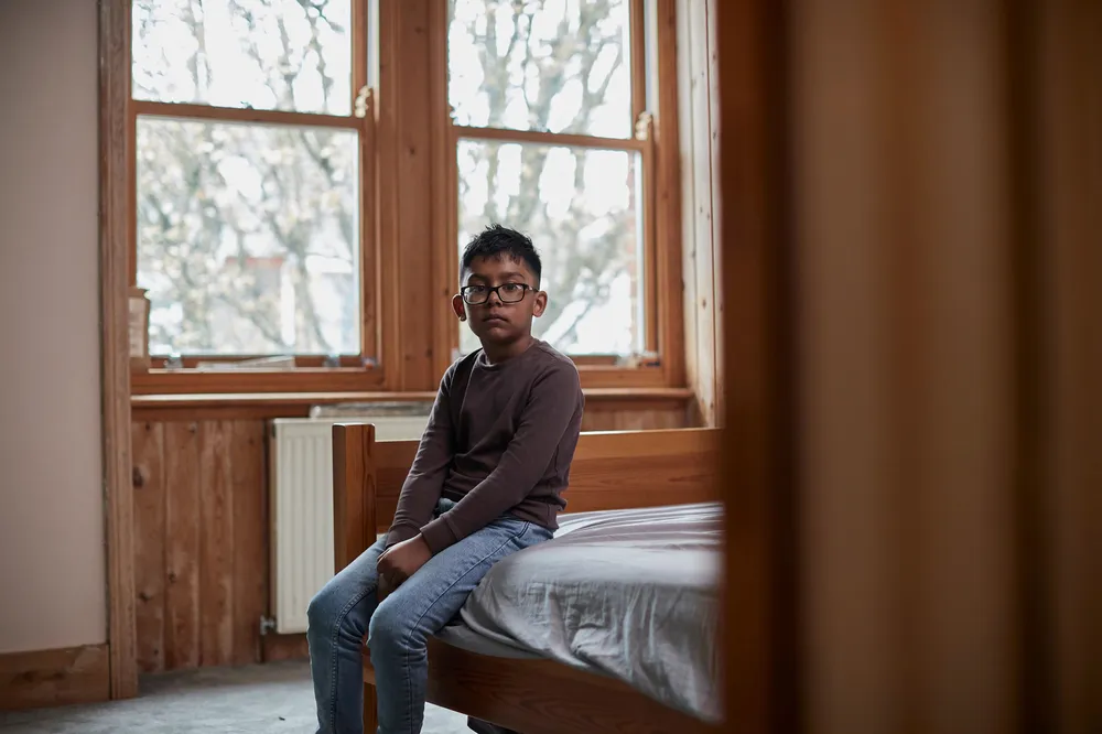 A young boy sitting on the edge of a wooden bed in a sparsely furnished room with large windows showing bare tree branches outside. The scene conveys a quiet, contemplative mood. The boy is dressed in a brown long-sleeve top and blue jeans.