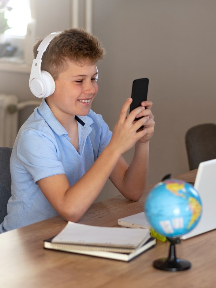 Young boy wearing headphones laughing at phone while sitting at a desk with an open laptop