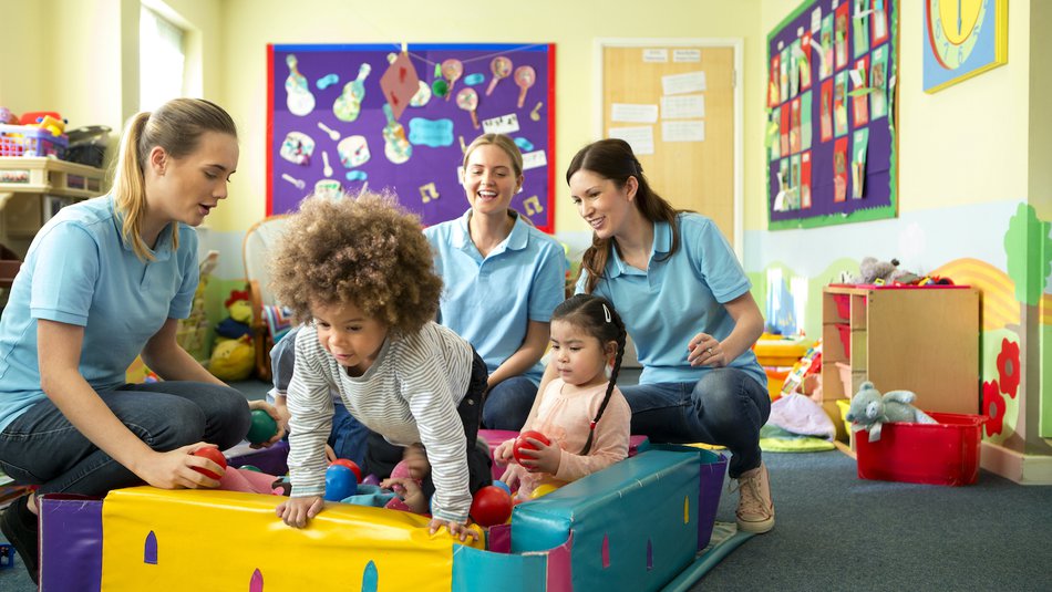 Young children playing in ball pit in nursery assisted by three workers