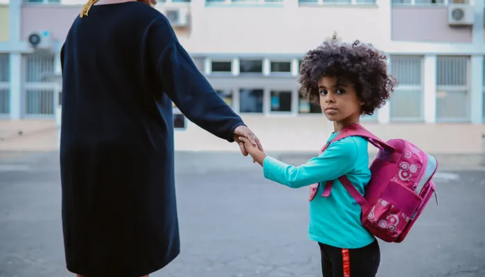 A child wearing a turquoise long-sleeve shirt and pink backpack holding hands with an adult dressed in a black coat, standing outside a school building. The scene suggests a moment of support, possibly a parent carer accompanying the child to school.