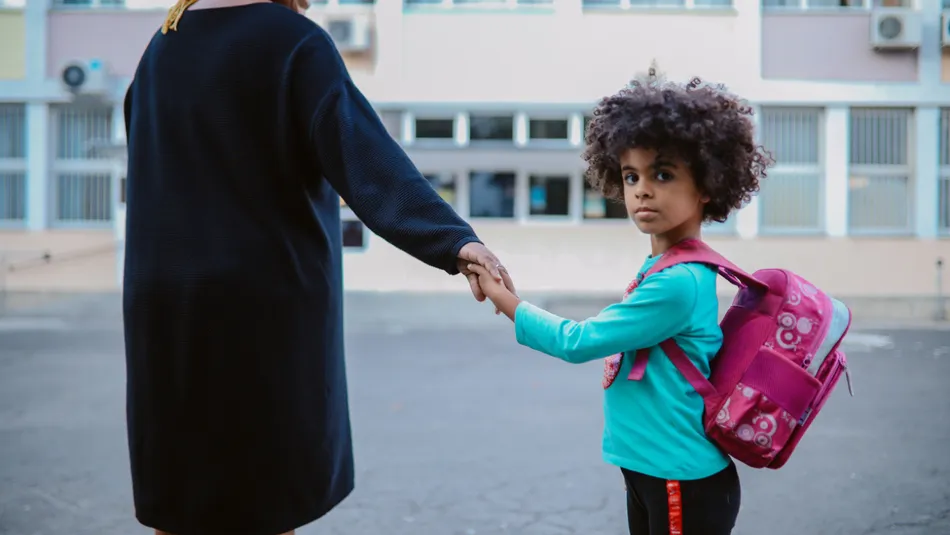 A child wearing a turquoise long-sleeve shirt and pink backpack holding hands with an adult dressed in a black coat, standing outside a school building. The scene suggests a moment of support, possibly a parent carer accompanying the child to school.