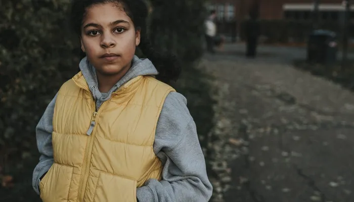 A person wearing a yellow gilet over a gray hoodie standing outdoors on a leaf-strewn pathway. Background includes blurred greenery and buildings, suggesting an urban park or residential area during autumn.