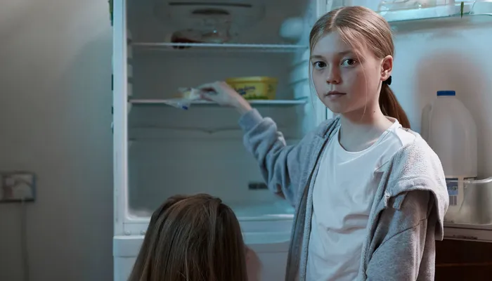 Two children standing in front of an open refrigerator in a kitchen. One child is reaching inside the fridge, which contains a few food items including a container of milk on the door and some cartons on the shelves.