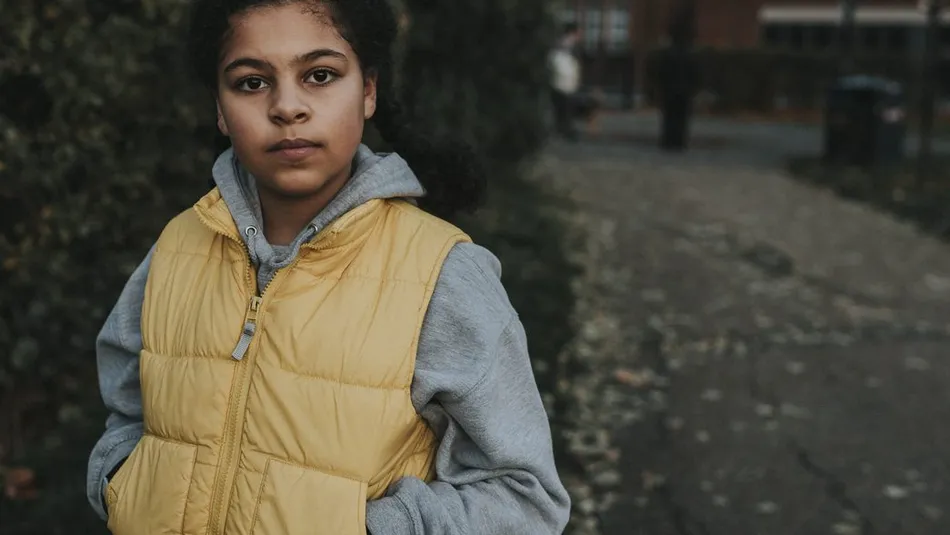 A person wearing a yellow gilet over a gray hoodie standing outdoors on a leaf-strewn pathway. Background includes blurred greenery and buildings, suggesting an urban park or residential area during autumn.