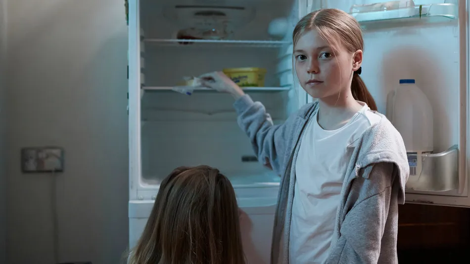 Two children standing in front of an open refrigerator in a kitchen. One child is reaching inside the fridge, which contains a few food items including a container of milk on the door and some cartons on the shelves.
