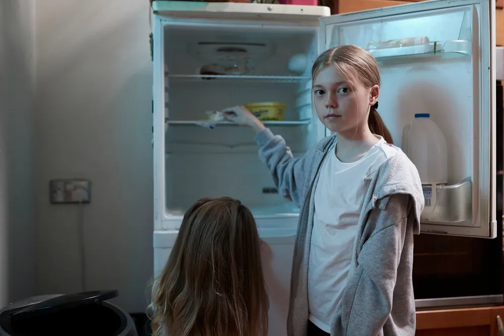 Two children standing in front of an open refrigerator in a kitchen. One child is reaching inside the fridge, which contains a few food items including a container of milk on the door and some cartons on the shelves.