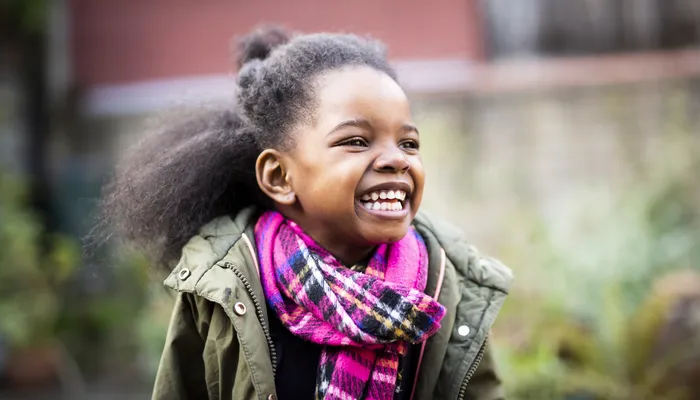 A smiling child wearing a green jacket and a vibrant pink and purple plaid scarf, standing outdoors with blurred background foliage and a brick wall. The image highlights colourful winter clothing, suggesting a cool weather setting.
