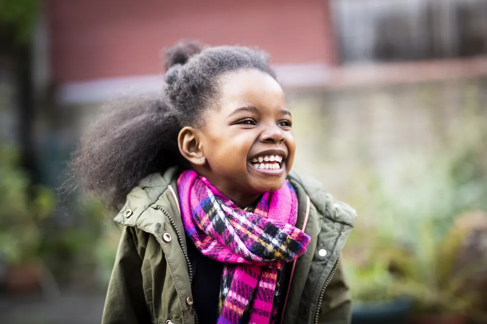 A smiling child wearing a green jacket and a vibrant pink and purple plaid scarf, standing outdoors with blurred background foliage and a brick wall. The image highlights colourful winter clothing, suggesting a cool weather setting.