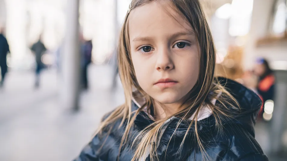 A young child with a serious expression wearing a black puffer jacket in an outdoor setting with blurred background featuring columns and people. The image captures casual urban atmosphere with natural lighting and soft focus on surroundings.