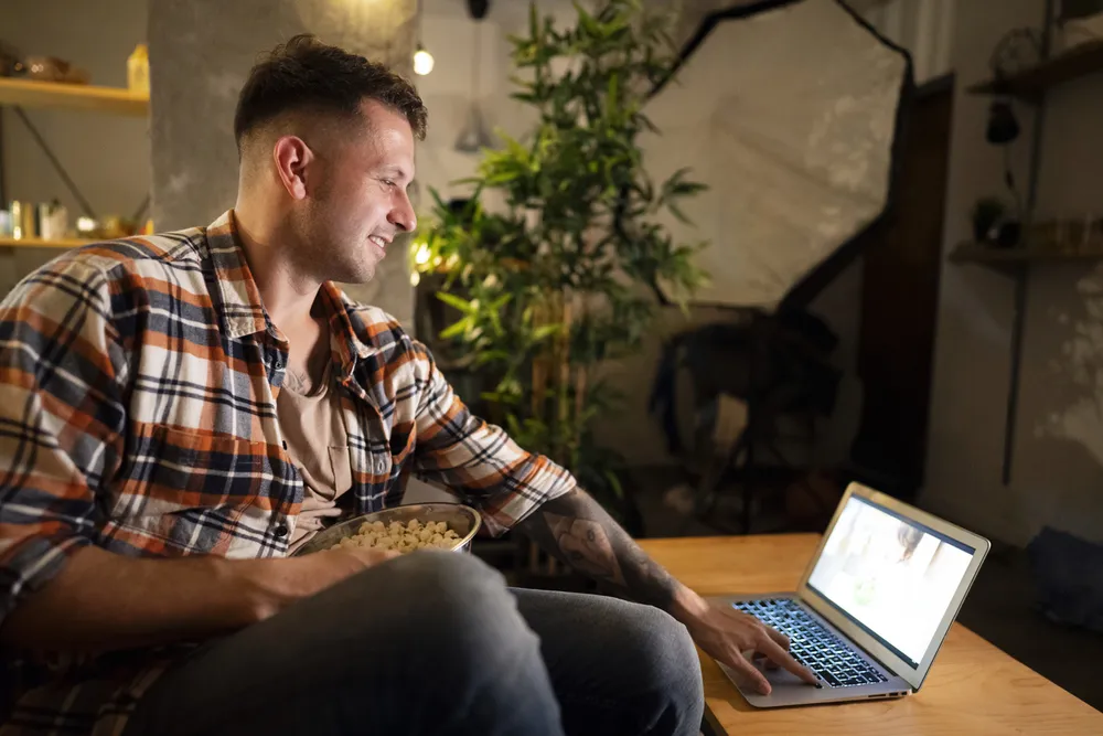 A person sitting in a cozy, dimly lit room, holding a bowl of popcorn and using a laptop on a wooden table. The setting includes indoor plants, shelves with decorative items, suggesting a relaxed environment for entertainment or work.