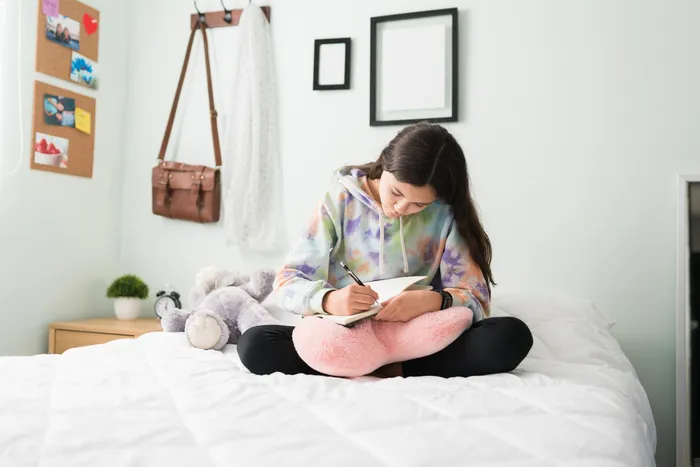 Young teen girl in her bedroom writing in a diary while sitting on her bed