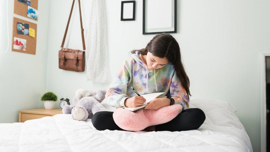 Young teen girl in her bedroom writing in a diary while sitting on her bed