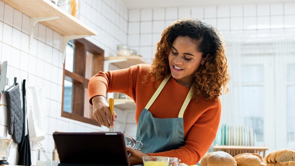 A person wearing an orange jumper and blue apron whisking ingredients in a bowl while looking at a tablet on a kitchen counter. Kitchen setting includes white tiled walls, wooden shelves with jars, and various baking items such as bread, eggs, and a lemon