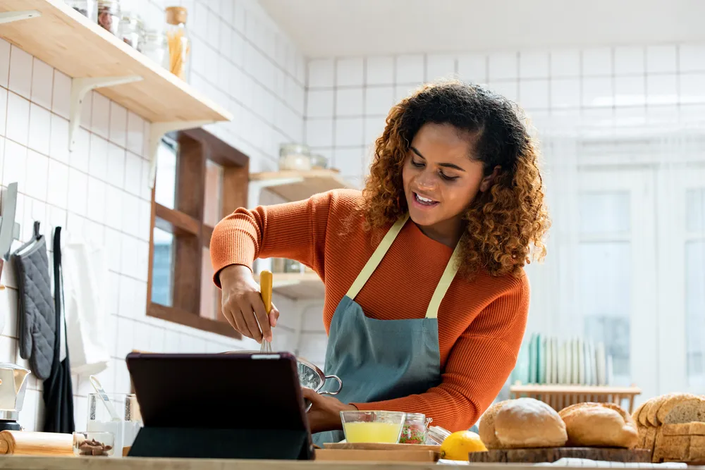 A person wearing an orange jumper and blue apron whisking ingredients in a bowl while looking at a tablet on a kitchen counter. Kitchen setting includes white tiled walls, wooden shelves with jars, and various baking items such as bread, eggs, and a lemon