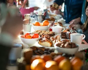 A festive breakfast or brunch table filled with various foods including bowls of fruit, pastries, cookies, and cups of coffee or tea. Several people are gathered around the table and serving themselves, creating a warm and social atmosphere
