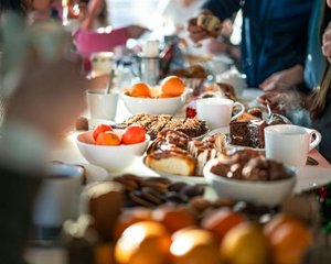 A festive breakfast or brunch table filled with various foods including bowls of fruit, pastries, cookies, and cups of coffee or tea. Several people are gathered around the table and serving themselves, creating a warm and social atmosphere