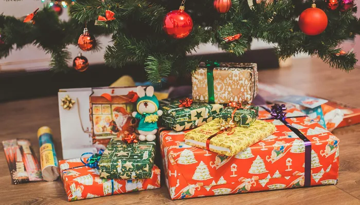 Colourful wrapped Christmas presents of various sizes arranged under a decorated Christmas tree with coloured baubles. The gifts feature festive wrapping paper with ribbons and bows and a small knitted toy sits among the packages on a wooden floor