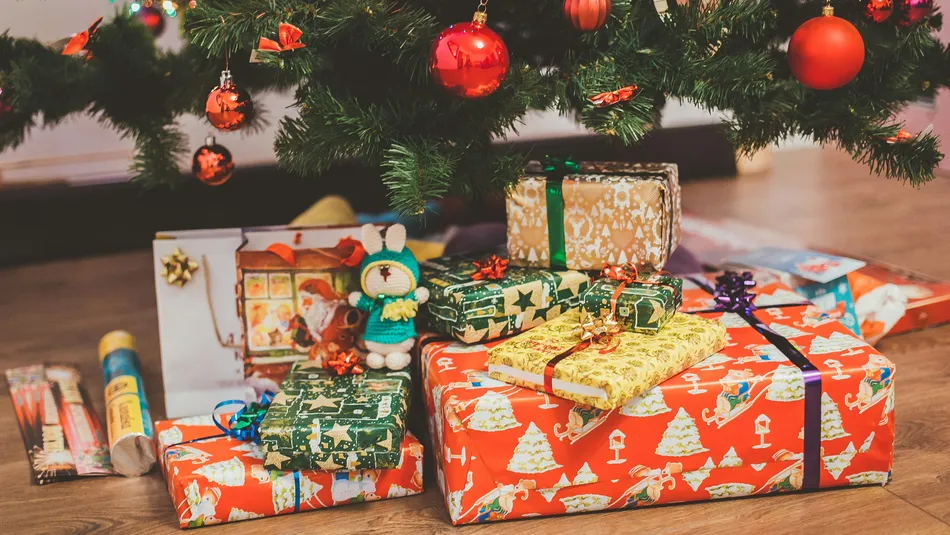 Colourful wrapped Christmas presents of various sizes arranged under a decorated Christmas tree with coloured baubles. The gifts feature festive wrapping paper with ribbons and bows and a small knitted toy sits among the packages on a wooden floor