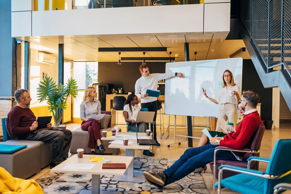 A modern office meeting with six diverse colleagues engaged in a presentation around a whiteboard. Bright room features casual seating, large windows, and a staircase, highlighting collaborative work environment and active discussion.
