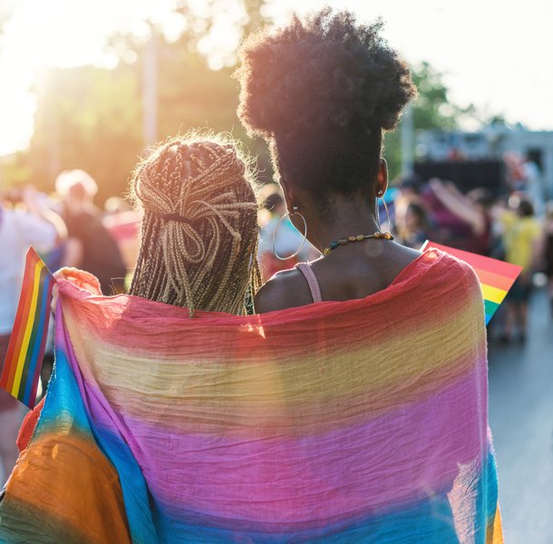 Two people walking in a pride parade, wrapped together in a rainbow flag symbolizing LGBTQ+ pride. The scene features sunlight filtering through trees, with other participants holding rainbow flags and celebrating in the background.