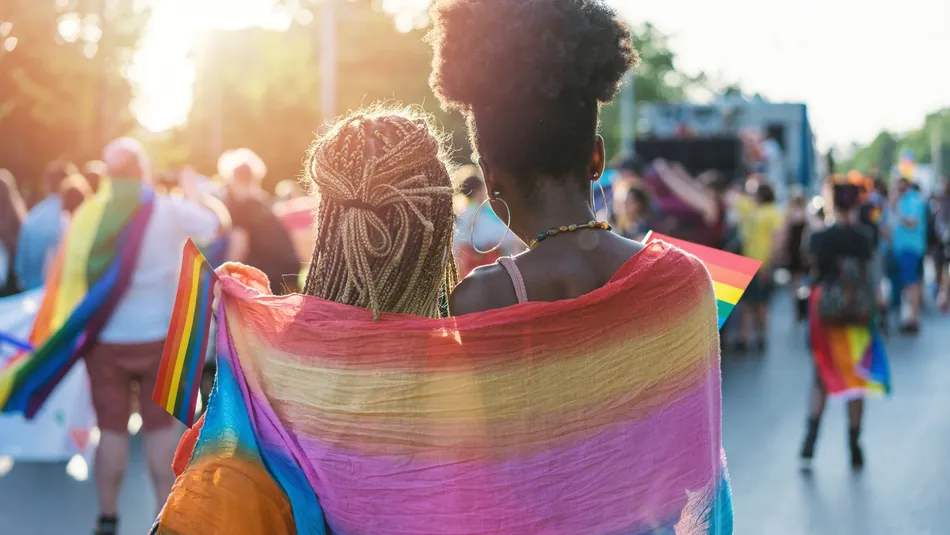 Two people walking in a pride parade, wrapped together in a rainbow flag symbolizing LGBTQ+ pride. The scene features sunlight filtering through trees, with other participants holding rainbow flags and celebrating in the background.