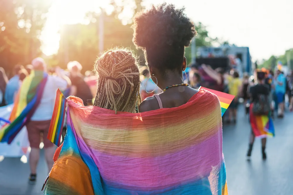 Two people walking in a pride parade, wrapped together in a rainbow flag symbolizing LGBTQ+ pride. The scene features sunlight filtering through trees, with other participants holding rainbow flags and celebrating in the background.
