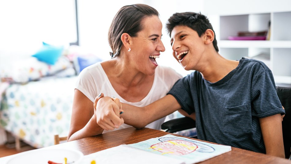 A woman and a boy sitting at a table engaged in a drawing activity, with the woman holding the boy's hand in a supportive gesture. The setting appears to be a bright room with shelves in the background.
