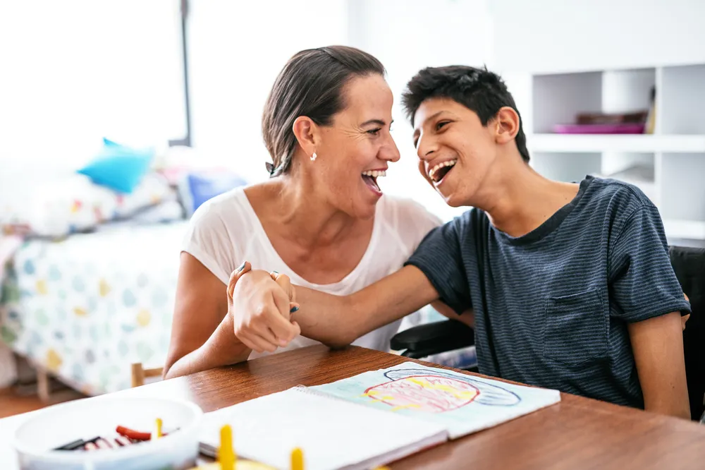 A woman and a boy sitting at a table engaged in a drawing activity, with the woman holding the boy's hand in a supportive gesture. The setting appears to be a bright room with shelves in the background.