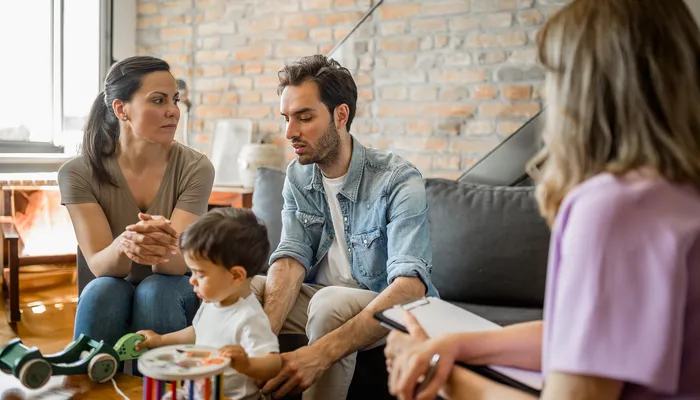 A family therapy session showing a couple and their child sitting on a couch while a therapist takes notes. The setting includes a cozy living room with toys on a table, highlighting a supportive environment focused on family communication and counselling