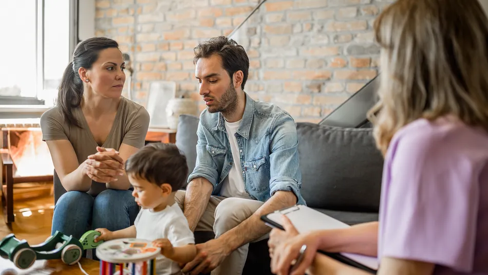 A family therapy session showing a couple and their child sitting on a couch while a therapist takes notes. The setting includes a cozy living room with toys on a table, highlighting a supportive environment focused on family communication and counselling