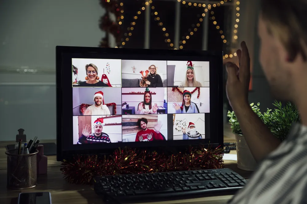 A computer screen showing a virtual holiday video call with nine participants wearing festive hats and sweaters, smiling and waving. The screen is decorated with Christmas lights and tinsel.