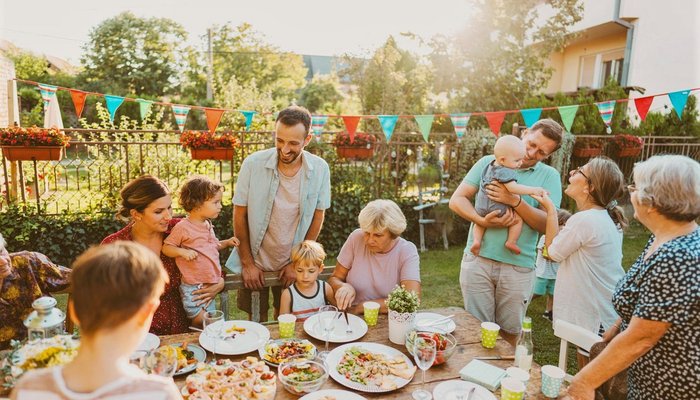 A multi-generational gathering outdoors around a table filled with various foods and drinks during a sunny day. Colourful triangular pennant banners hang in the background, people are interacting, including a man holding a baby.